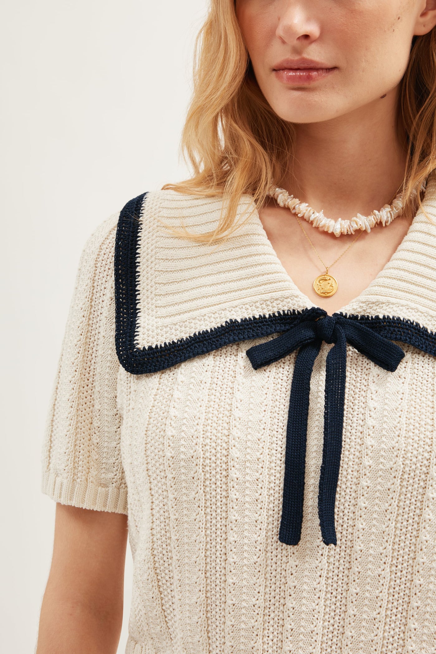 A woman wears the Pippa - Ivory, a cream summer knit top featuring a large textured sailor collar with navy trim and bow. She has blonde hair, puffed sleeves, and accessorizes with a shell and gold pendant necklace.