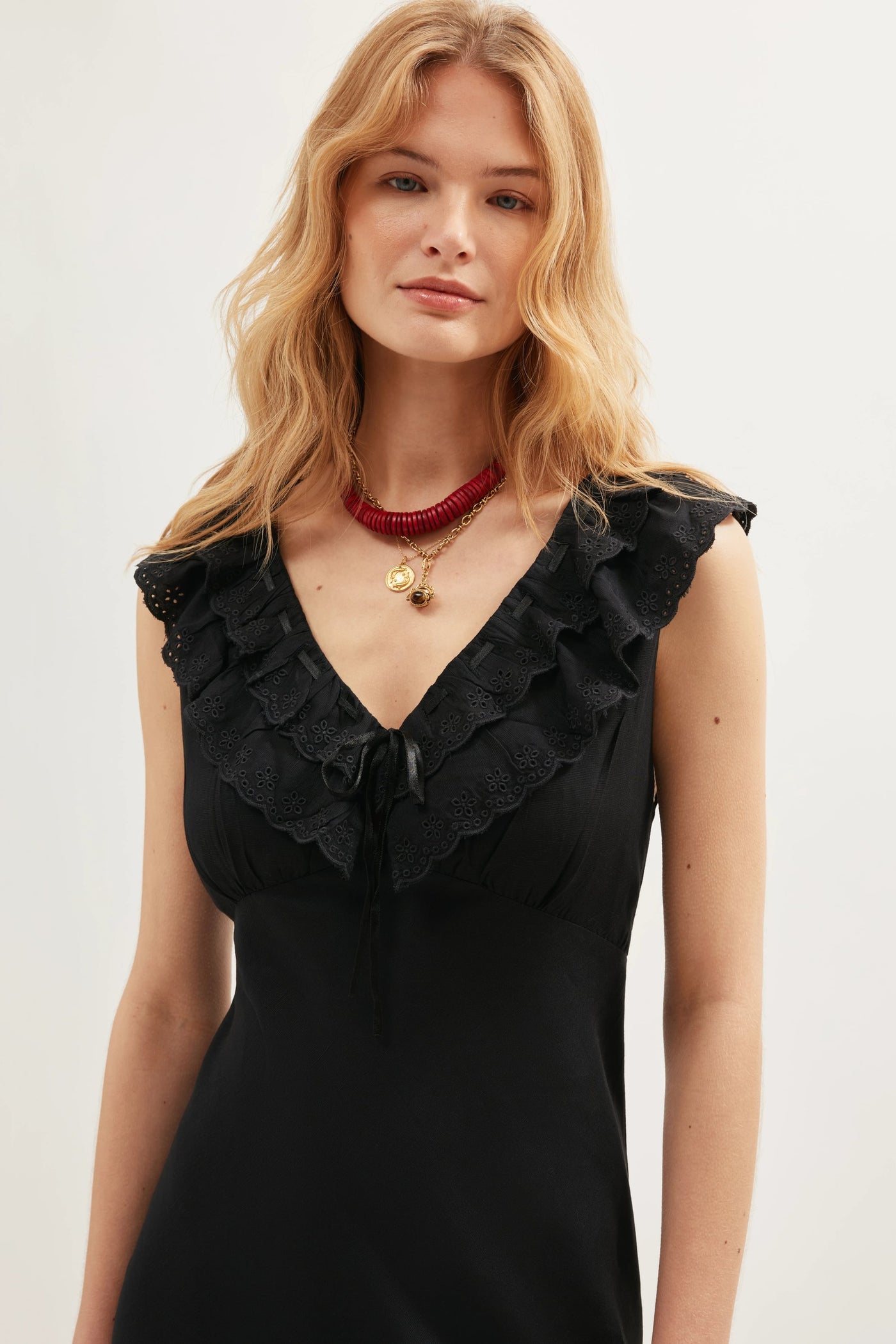 A woman with wavy blonde hair wears the sleeveless Seraphina - Black dress, showcasing a ruffled eyelet-trimmed V-neckline and scalloped ruffles. She accessorizes with layered necklaces: a red beaded choker and a gold pendant.