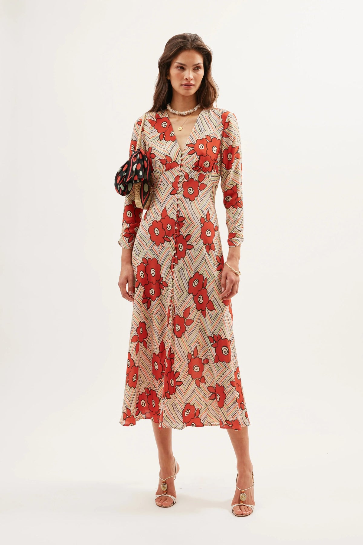 A woman wears the Katie - Milos Floral Cream dress, a long-sleeved, ankle-length piece with bold red floral graphics, holding a matching bag and wearing strappy heels against a plain white background.