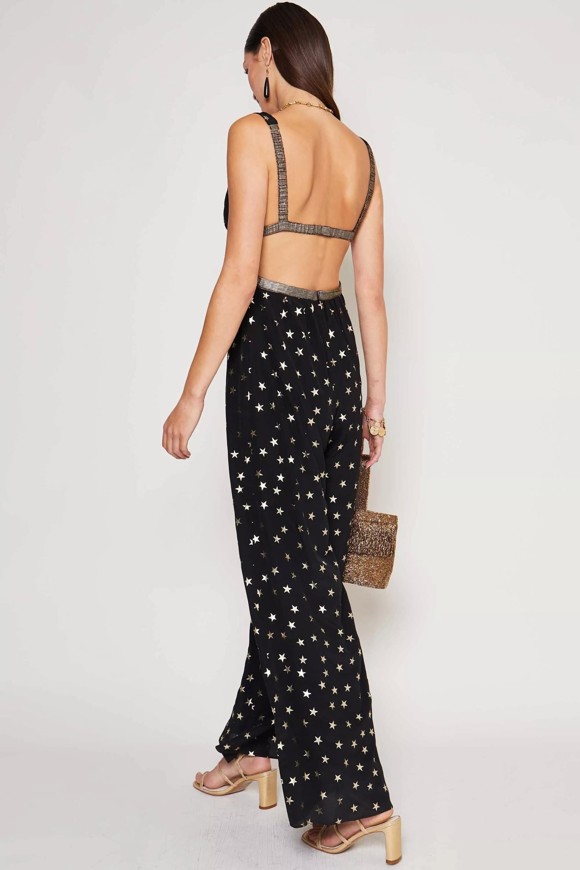 A woman faces away, party ready in the Hollywood - Star Foil black sleeveless jumpsuit with gold star print, holding a textured handbag and wearing strappy heels, earrings, and bracelets.