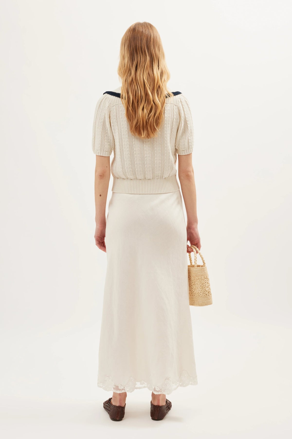 A woman with long, wavy blonde hair, seen from behind, wears the Pippa - Ivory: a vintage-style top with puffed sleeves and a white, lace-trimmed ankle skirt. She holds a woven bag and wears dark shoes against a plain white background.