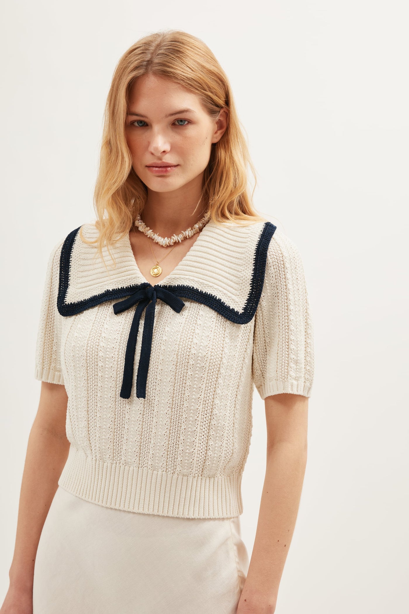 A blonde woman wears the Pippa - Ivory vintage-style cream knit top, featuring short sleeves, a wide collar with dark trim, and a front bow. She pairs it with a necklace and light skirt against a plain background.