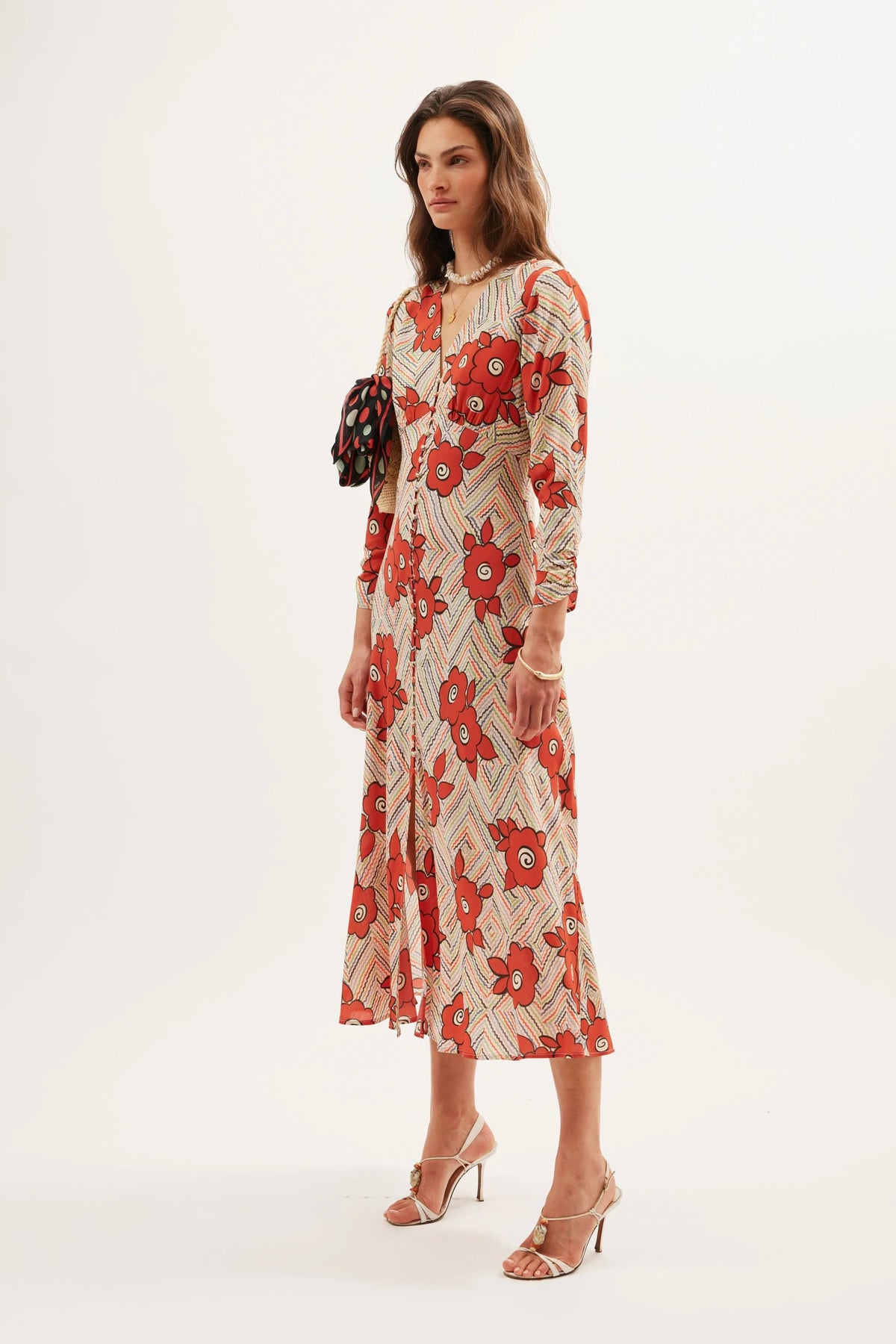 A woman stands against a white background wearing the Katie - Milos Floral Cream dress with red flowers and beige zigzag lines, metallic heeled sandals, and carrying a small black handbag with colorful details.