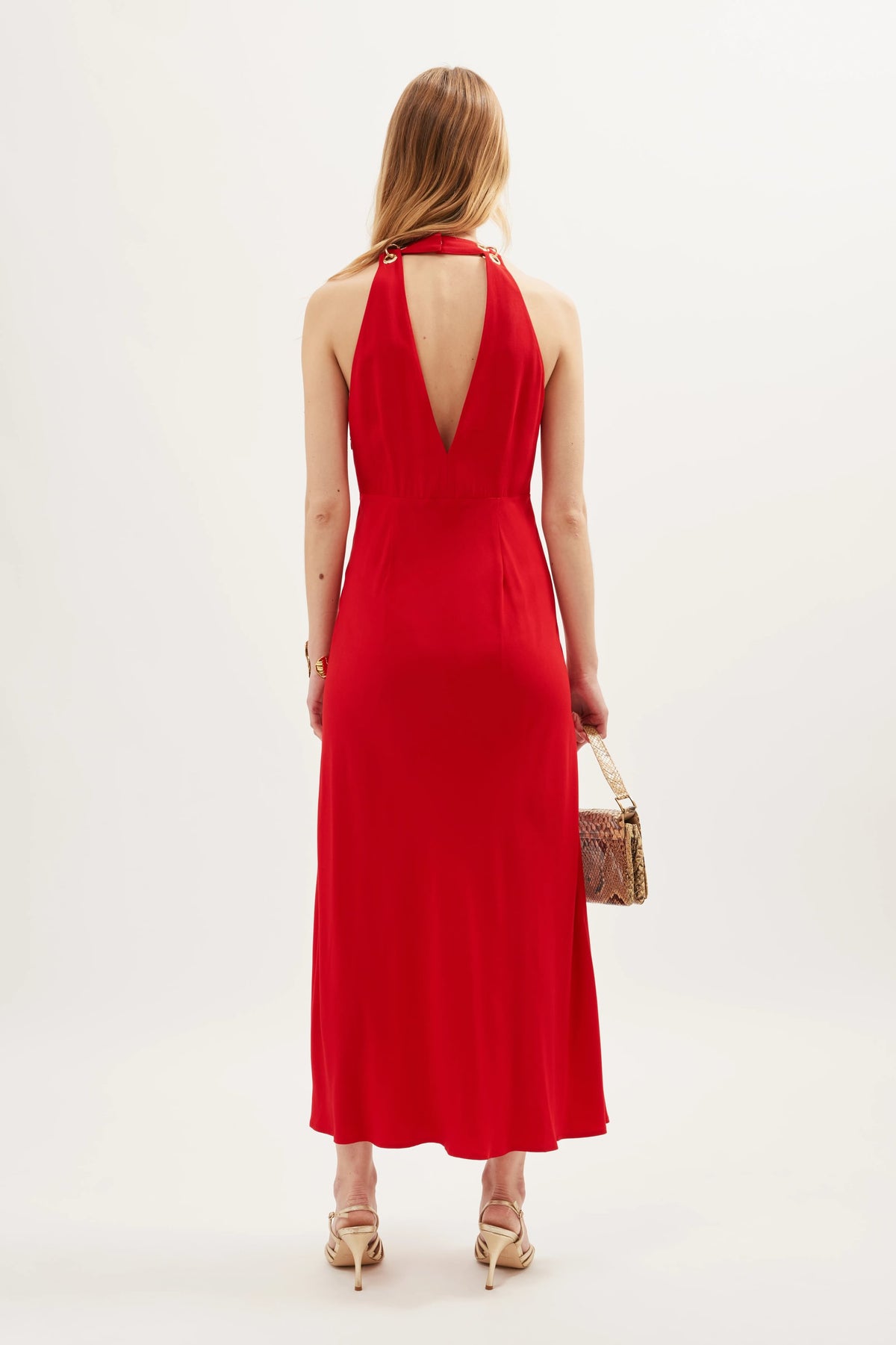 A woman stands facing away, wearing the Eleanora - Red dress—a sleeveless, vintage-inspired design featuring a deep V-back cutout. She holds a small brown handbag and wears nude heels. Her long blonde hair is loose, set against a plain white background.
