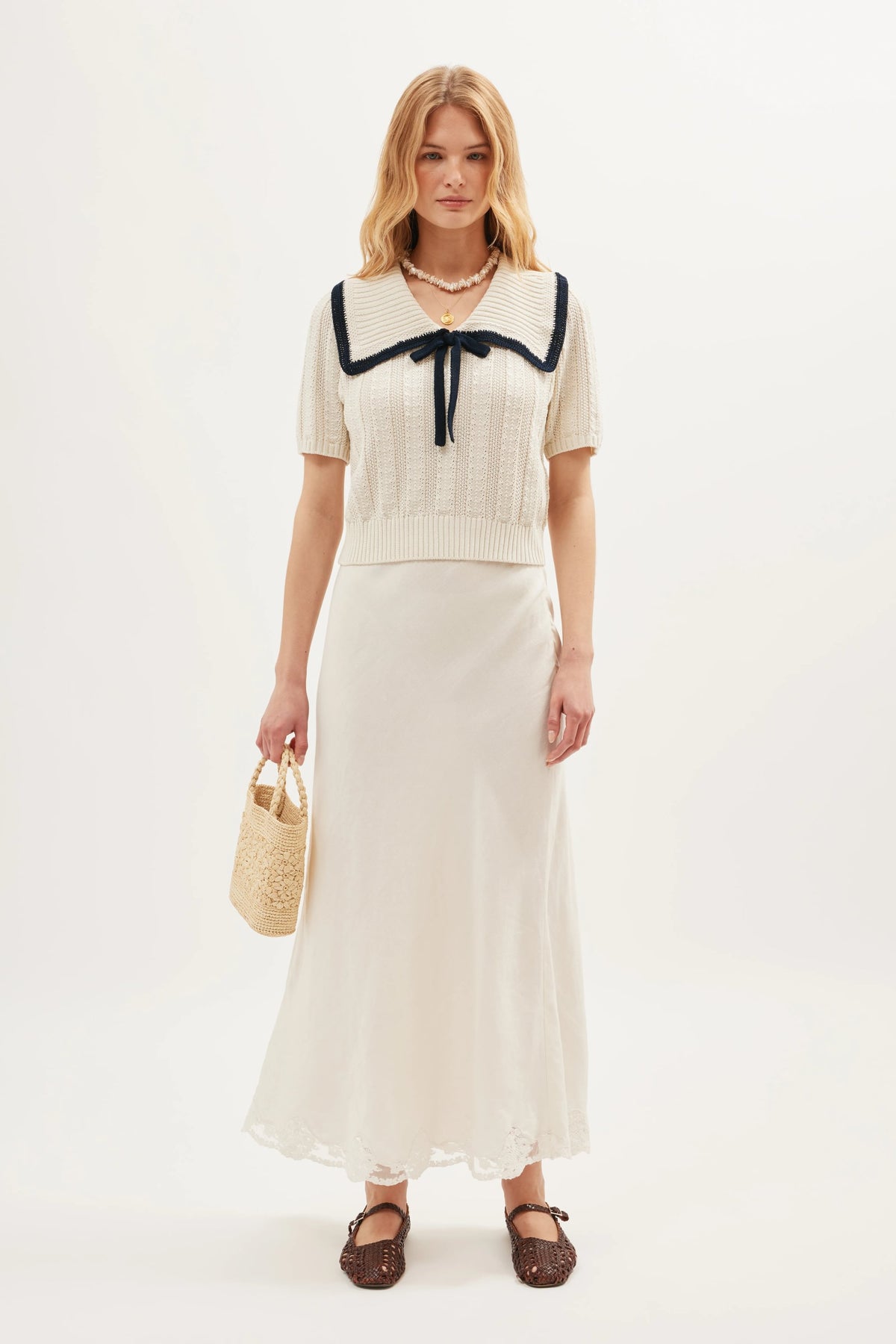 A woman stands against a plain light background wearing the Pippa - Ivory top, a vintage-style cream nautical knit with a wide black-trimmed sailor collar, paired with a long cream skirt, brown shoes, and a woven handbag.
