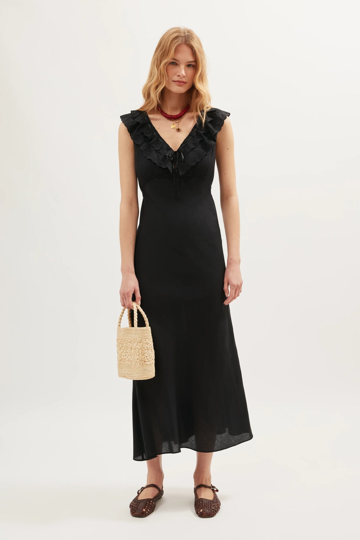 A woman wears the Seraphina - Black dress, a sleeveless style with scalloped ruffles along its V-neckline. She stands against a plain background, accessorizing with a woven handbag, brown shoes, and layered necklaces.
