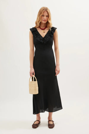 A woman wears the Seraphina - Black dress, a sleeveless style with scalloped ruffles along its V-neckline. She stands against a plain background, accessorizing with a woven handbag, brown shoes, and layered necklaces.