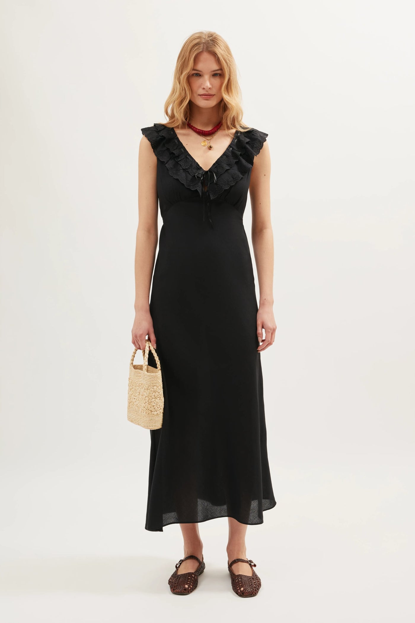 A woman stands against a plain background wearing the Seraphina - Black, a sleeveless midi dress with scalloped ruffles along the V-neckline. She accessorizes with a straw handbag, brown woven ankle-strap shoes, and a dark choker necklace.