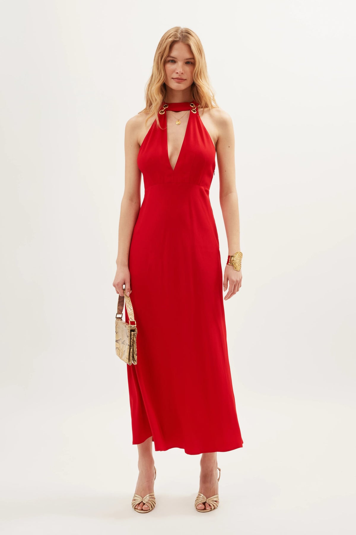 Wearing the Eleanora - Red, a sleeveless vintage-inspired halter-neck dress featuring a deep V-neckline, she holds a gold handbag and accessorizes with gold heels and a bracelet, standing against a plain light background.