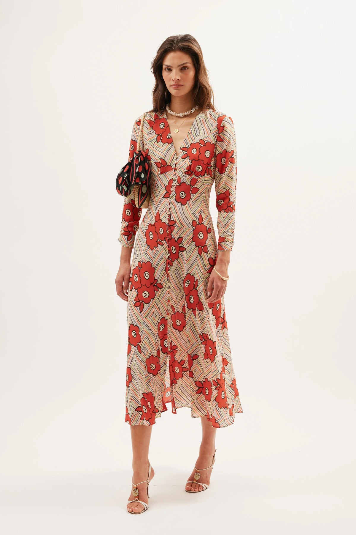 A woman poses in the Katie - Milos Floral Cream dress featuring long sleeves and a red-beige floral print. She holds a black handbag with circular accents, wears strappy sandals, gold jewelry, and stands against a plain white background.