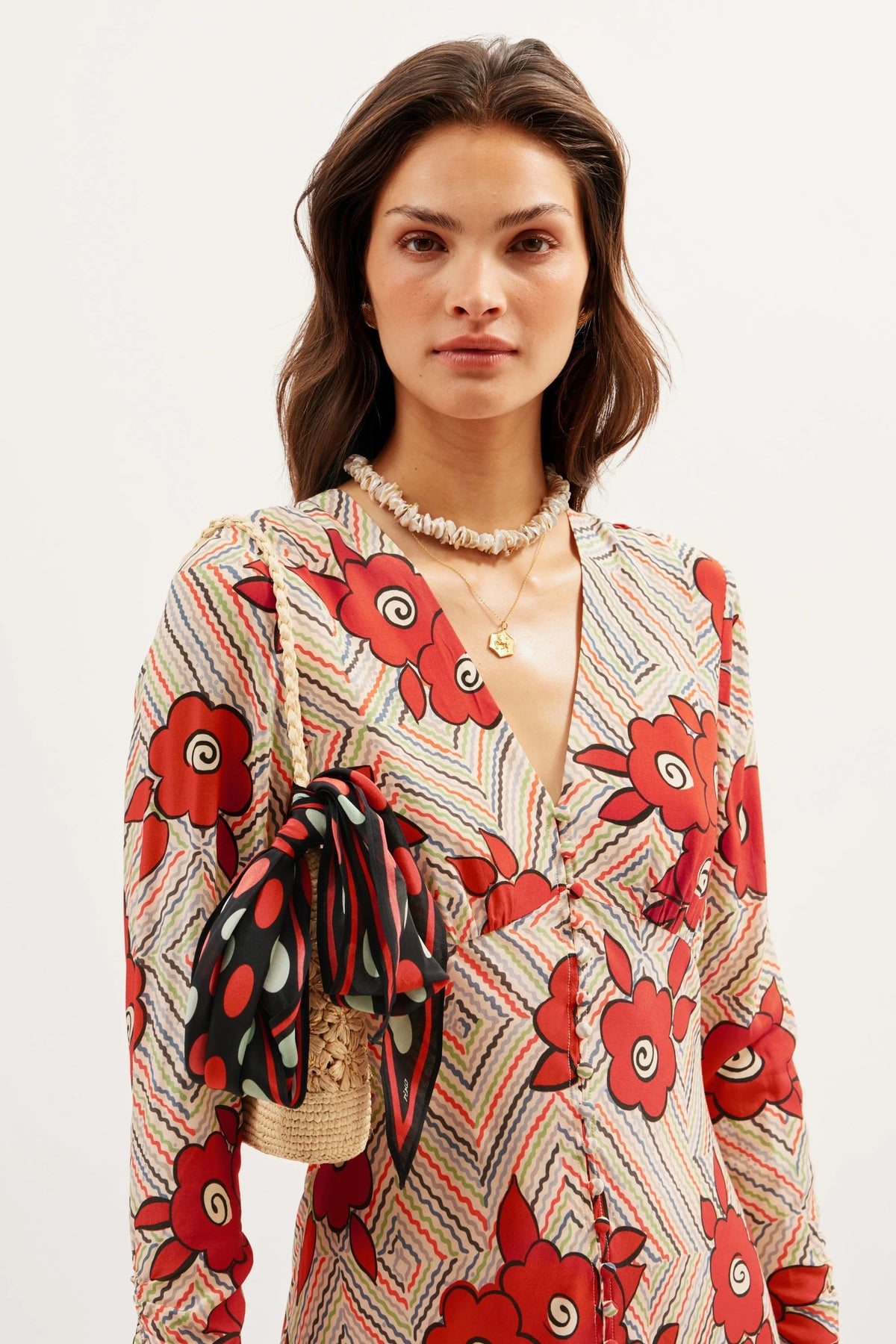 A woman in the Katie - Milos Floral Cream V-neck dress with a bold red floral and geometric print holds a woven bag with a polka-dot scarf. Her loose, wavy brown hair is styled with layered necklaces.