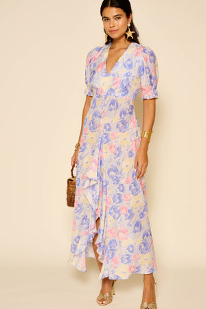 A woman wears the Ariel - Tulip Fields Blue vintage-inspired dress with short puff sleeves and a ruffled front hem, styled with gold jewelry, star-shaped earrings, and a small woven handbag against a beige background.