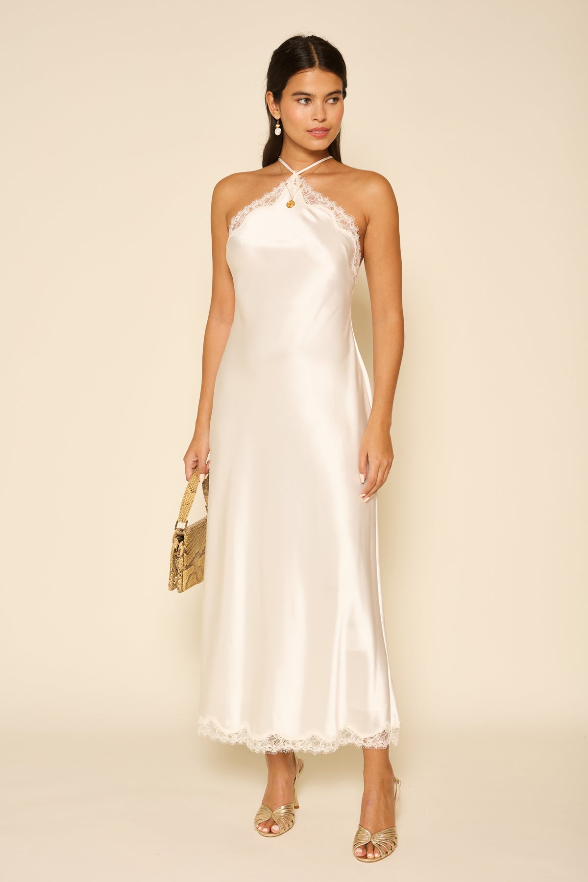 A woman poses against a beige background in the Harlan - Ivory, an ivory satin slip dress with lace trim. She accessorizes with gold earrings, a small gold handbag, and gold open-toe heels—perfect for special occasions.