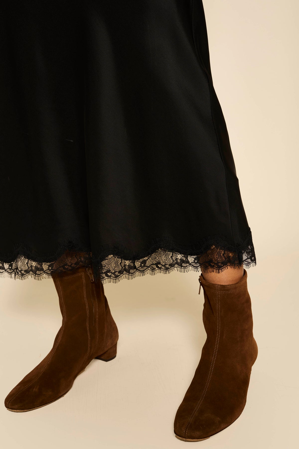 A person wearing a long black skirt with lace trim at the hem and Rey - Chocolate brown suede ankle boots stands on a light-colored floor.
