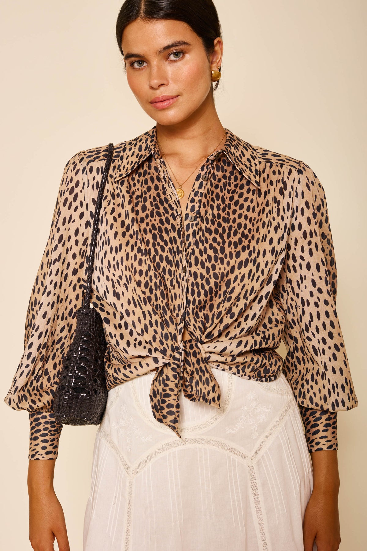 A woman wears the Blake - Antelope Spot vintage animal print shirt tied at the waist, paired with a white lace skirt, gold earrings, a necklace, and a black textured shoulder bag against a neutral background.