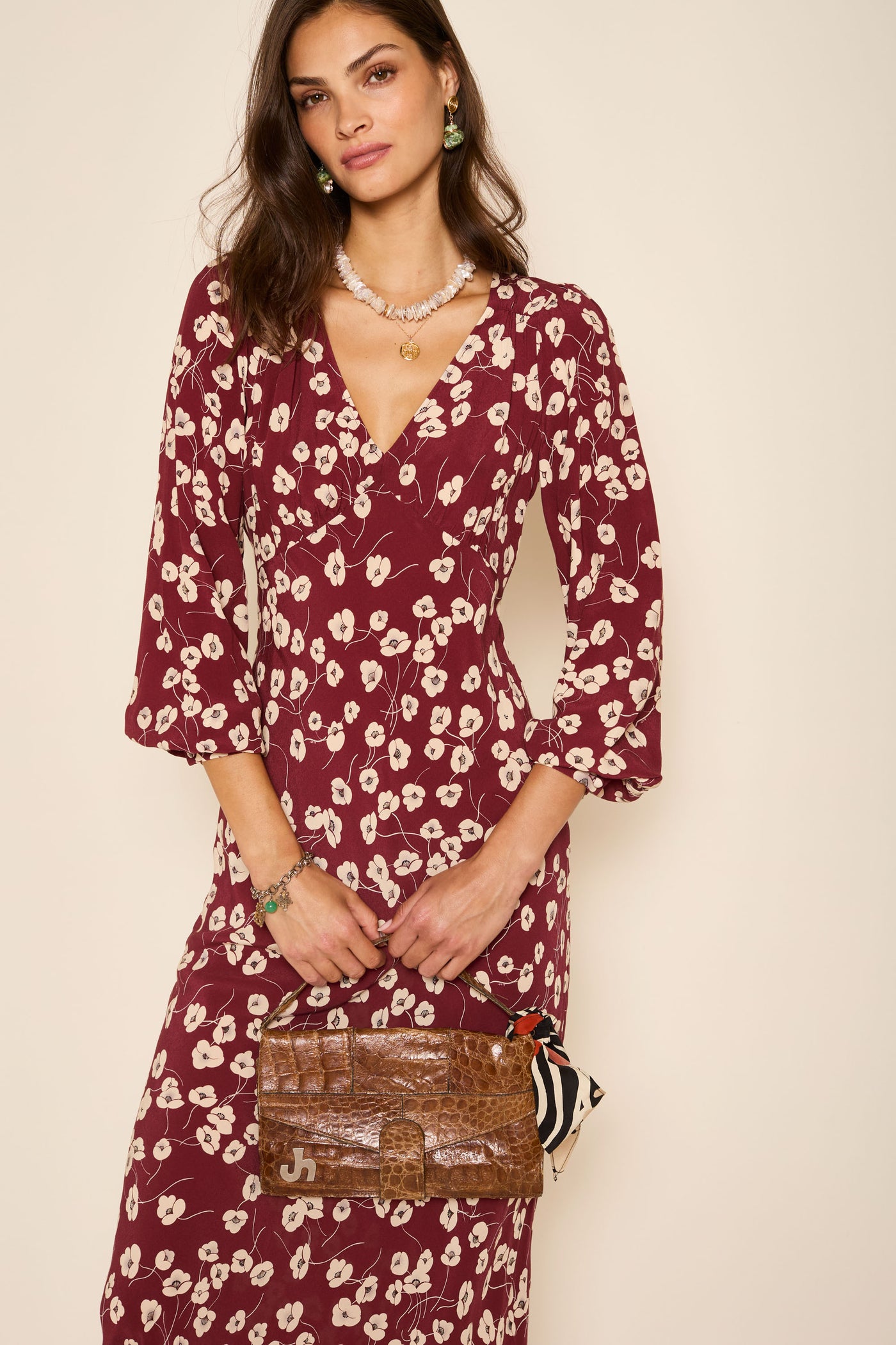A woman in the Mavis - Autumn Cherry Maroon long-sleeve dress with everyday florals stands against a beige background, holding a brown textured handbag with a scarf. She has loose brown hair and wears jewelry.