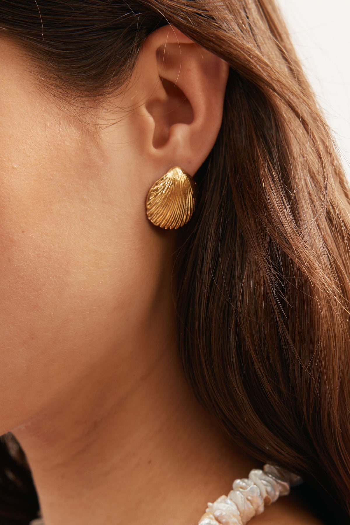 Close-up of a woman's ear wearing Meyra - Gold shell-shaped earrings. Her brown hair is tucked behind her ear, and she pairs them with a beaded necklace. The softly blurred background adds a delicate touch.