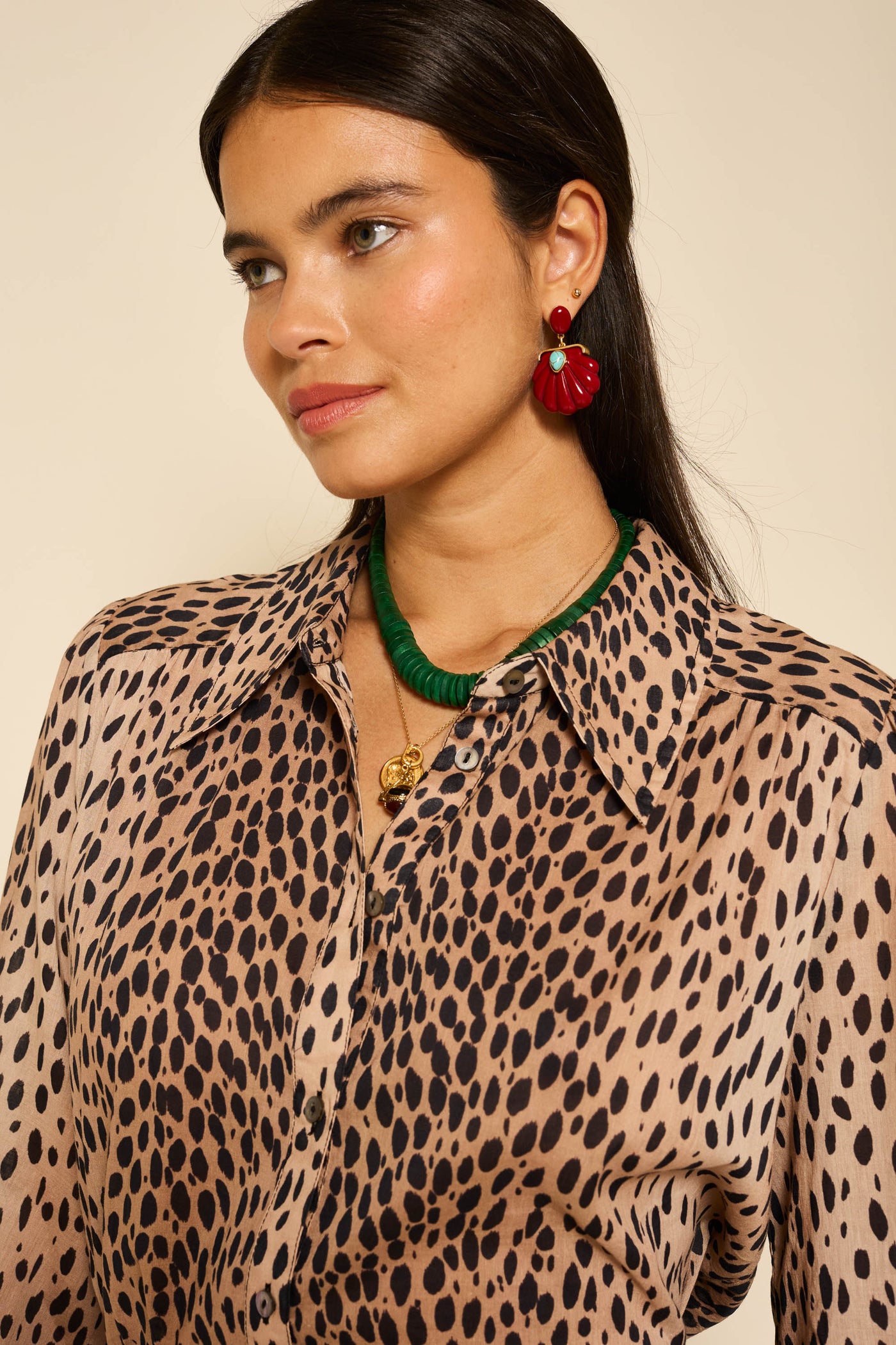 A woman with long dark hair models the Blake - Antelope Spot animal print vintage shirt, paired with a green beaded necklace, gold pendant, and red statement earrings, glancing sideways against a neutral background.