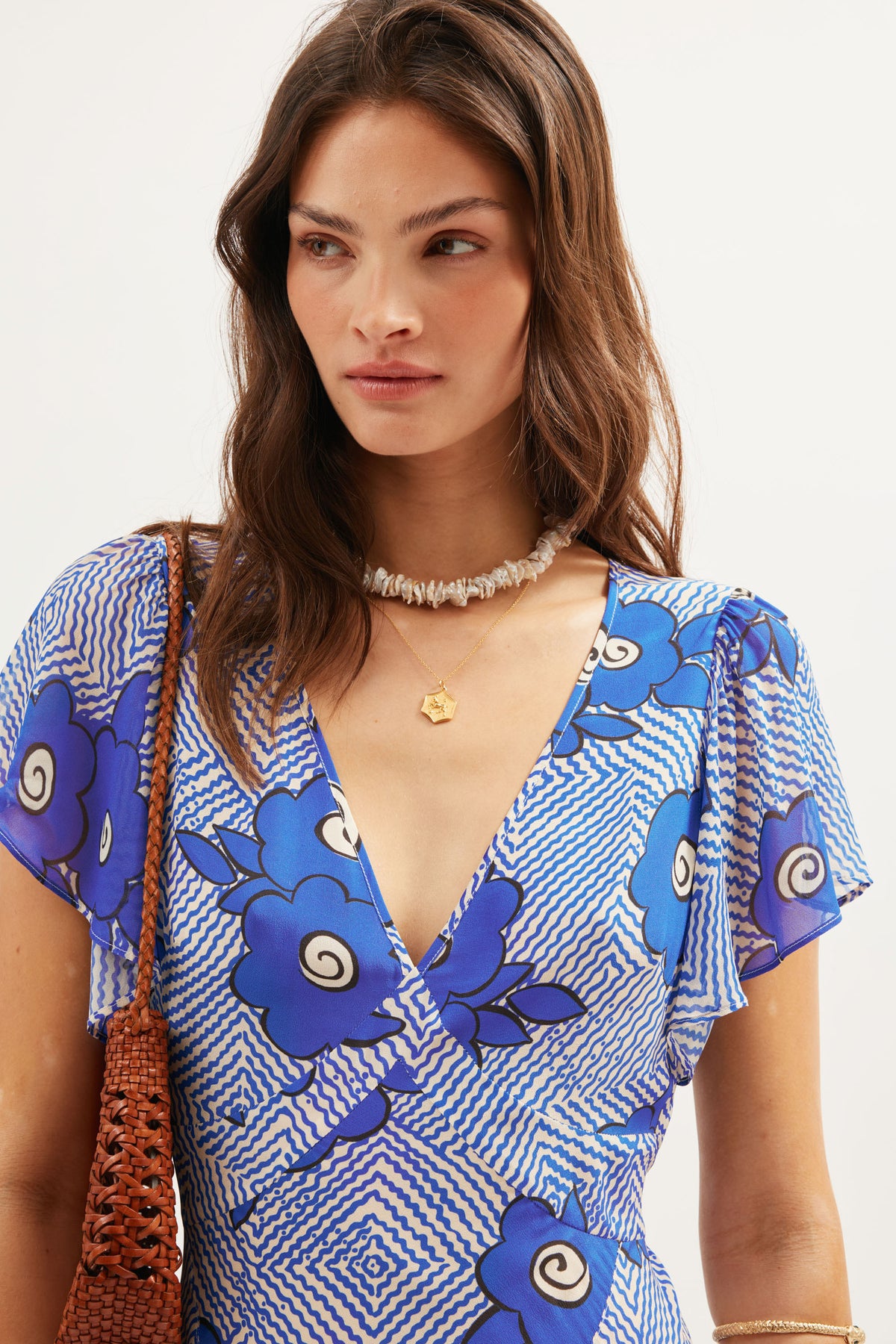 A woman with wavy brown hair wears the Evie - Milos Floral Blue dress, featuring bold florals and georgette ruffles. She accessorizes with a shell necklace, gold pendant, and a brown woven bag, standing against a plain white background.