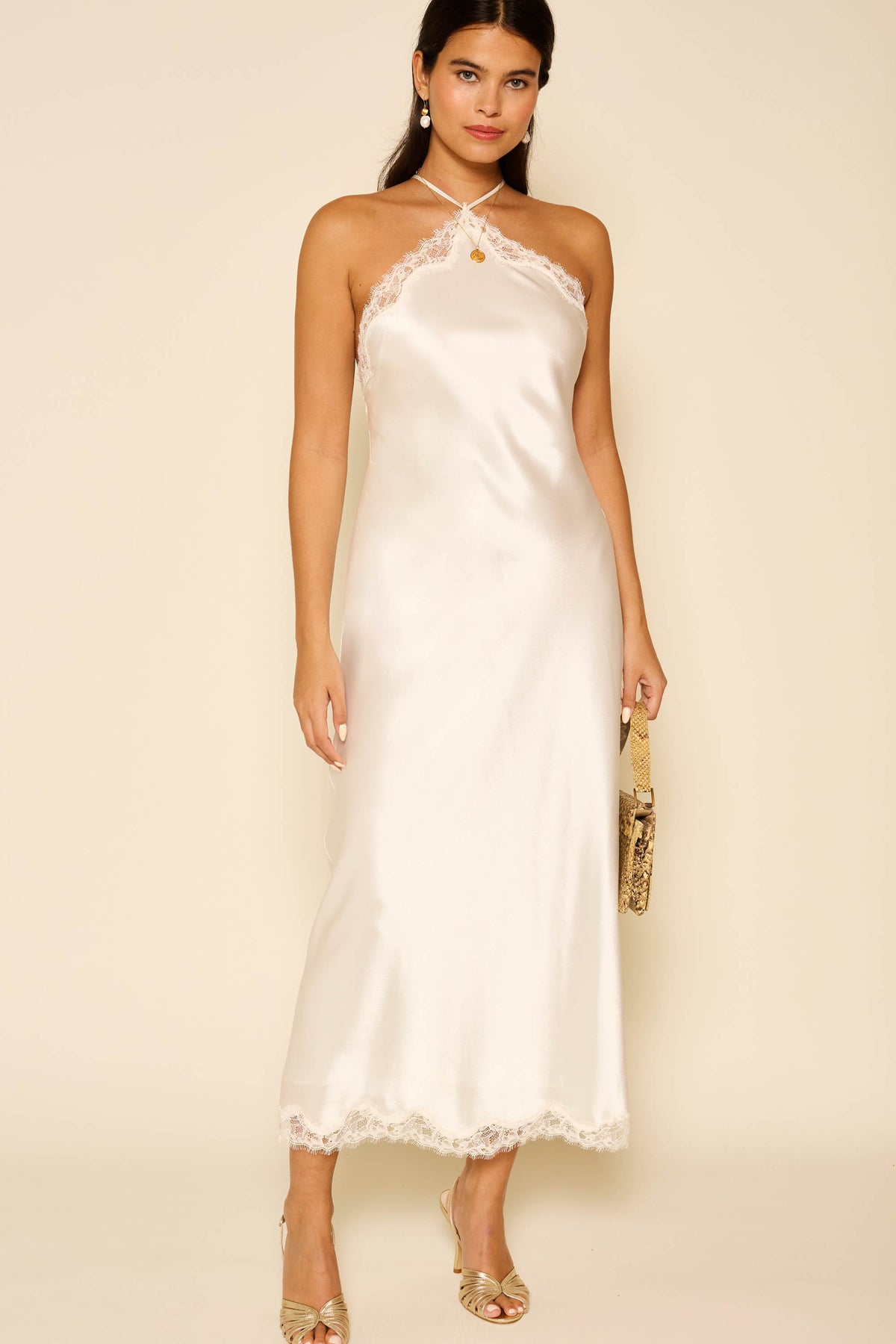 Woman in the Harlan - Ivory: a sleeveless satin slip dress with lace trim, strappy neckline, and midi length—an elegant pick for special occasions. She holds a clutch and wears open-toe heels, set against a neutral background.