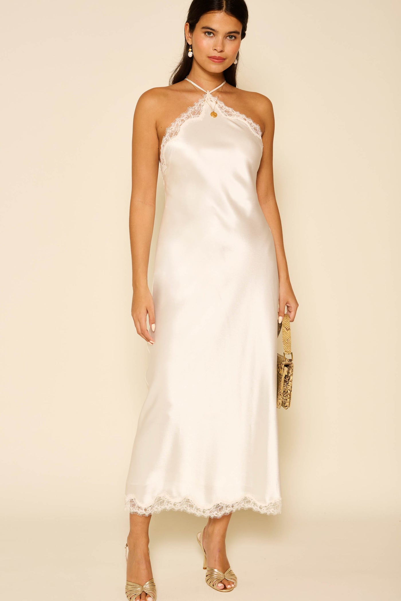 Woman in the Harlan - Ivory: a sleeveless satin slip dress with lace trim, strappy neckline, and midi length—an elegant pick for special occasions. She holds a clutch and wears open-toe heels, set against a neutral background.
