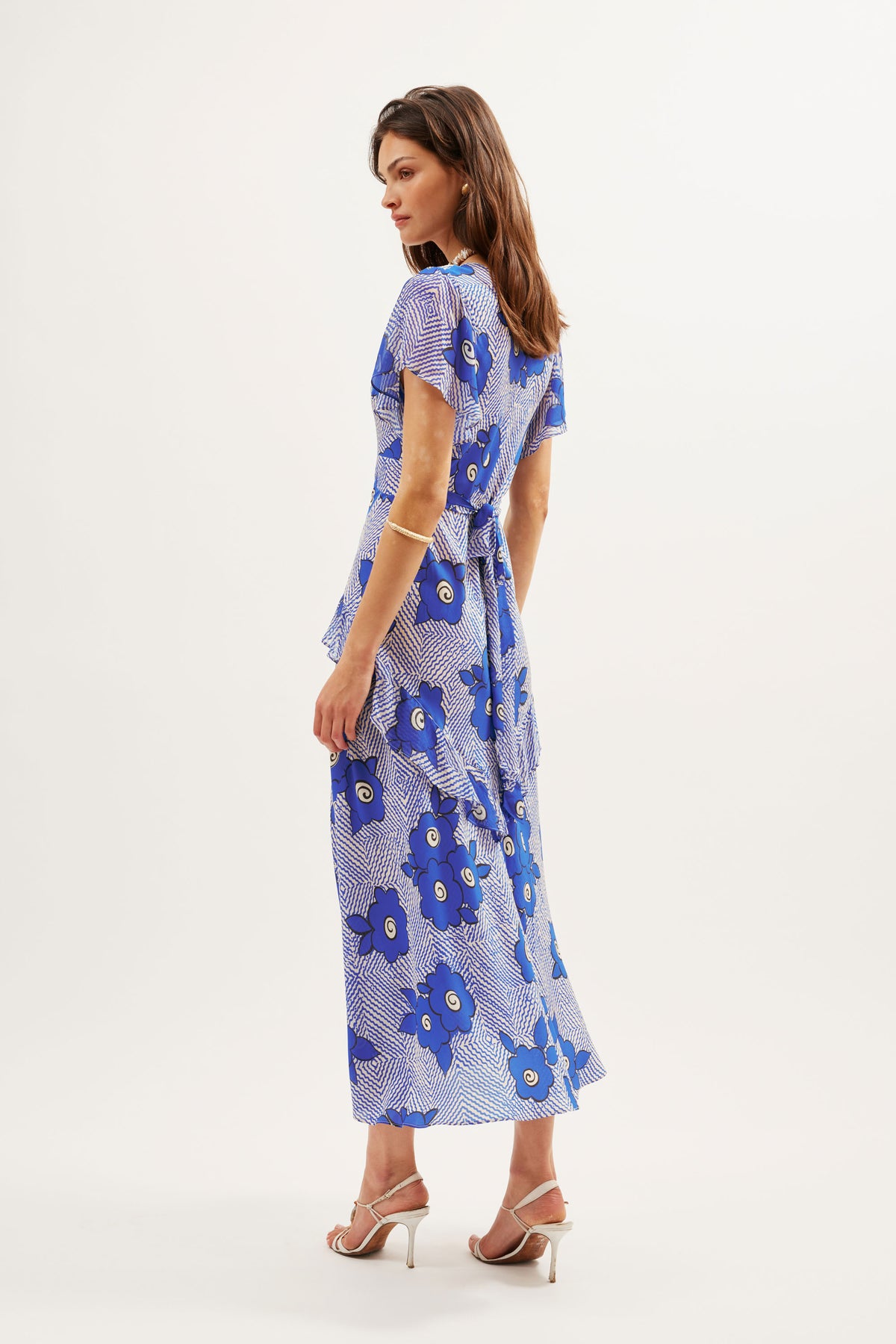 A woman stands sideways wearing the Evie - Milos Floral Blue, an ankle-length floral wrap dress in blue silk crepe de chine with short sleeves and white high-heeled sandals, set against a plain white background.