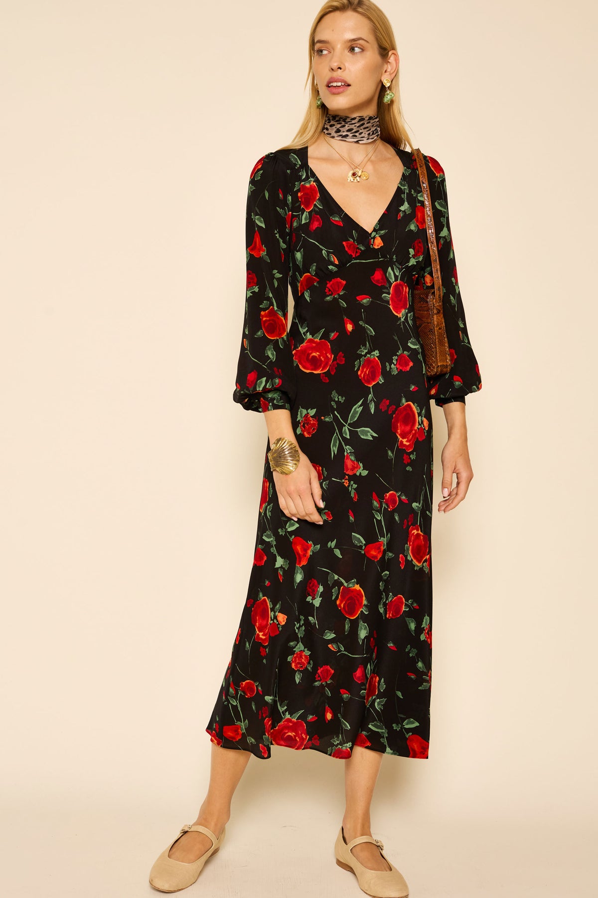 A woman in the Mavis - Claudia Rose Black dress, beige flats, a brown shoulder bag, a black patterned choker, and gold jewelry stands against a neutral background.