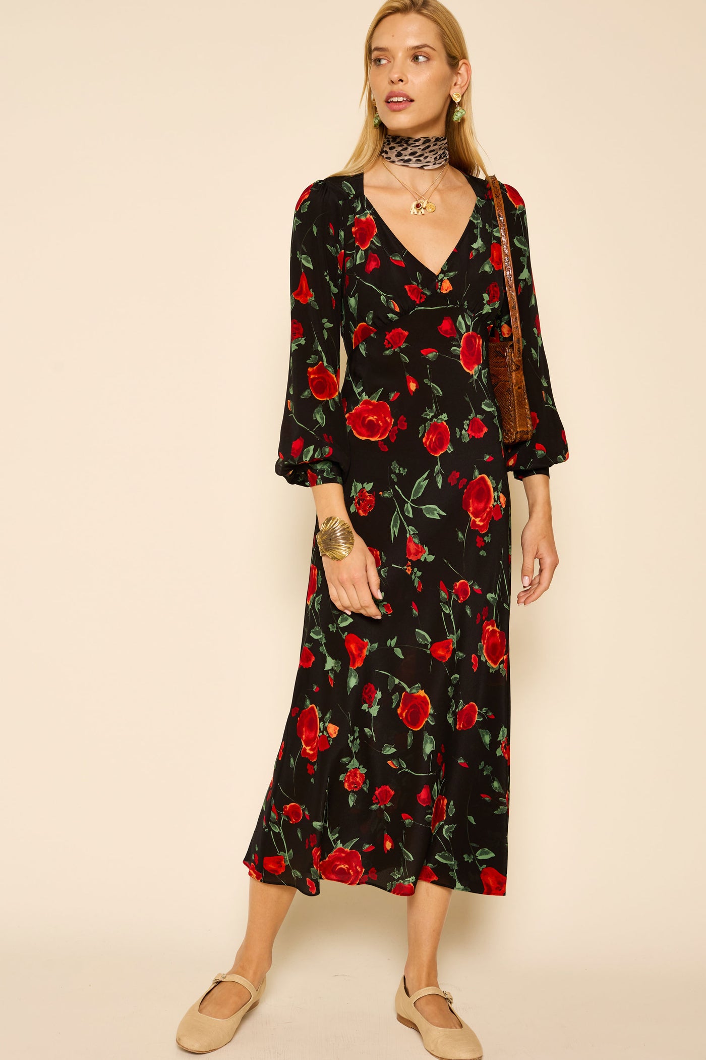 A woman in the Mavis - Claudia Rose Black dress, beige flats, a brown shoulder bag, a black patterned choker, and gold jewelry stands against a neutral background.