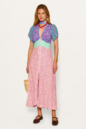 A woman wears the Novara - Shell Pansy Mix Multi vintage dress with curved sleeves, fitted waist, and front buttons, styled with a scarf, gold bracelet, woven bag, and brown shoes.