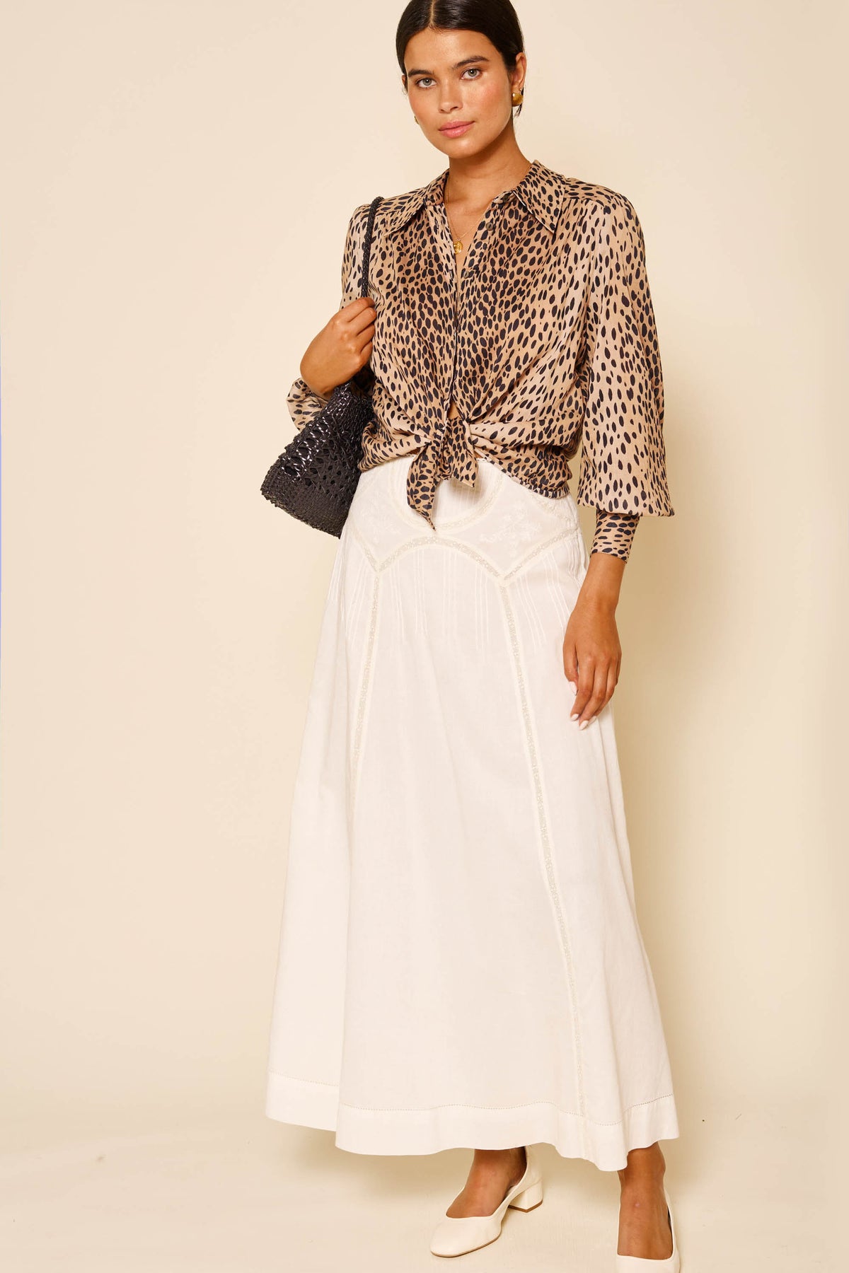 A woman stands against a beige background wearing the Vienne - Pansy Embroidery Ivory vintage-inspired white skirt, paired with a leopard-print blouse, cream shoes, and a black woven shoulder bag.