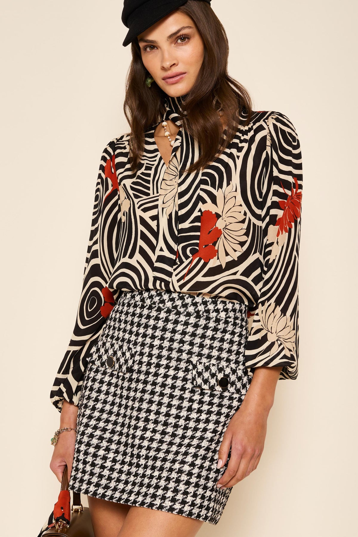 A woman wearing the Mariah - Houndstooth Mono mini skirt, a black beret, a beige, black, and red patterned blouse, and holding a brown handbag stands against a beige background for a noughties-inspired look.