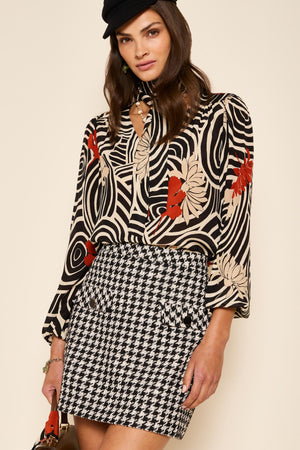 A woman wearing the Mariah - Houndstooth Mono mini skirt, a black beret, a beige, black, and red patterned blouse, and holding a brown handbag stands against a beige background for a noughties-inspired look.