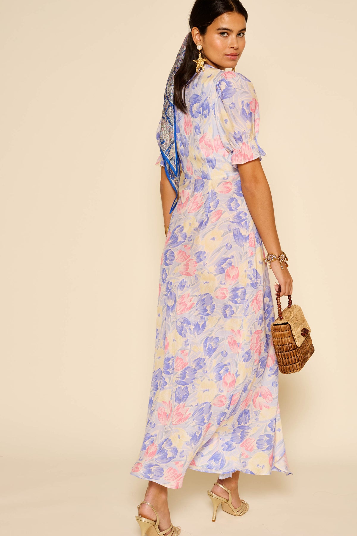 A woman wears the Ariel - Tulip Fields Blue dress with puff sleeves, facing backward and glancing over her shoulder. She accessorizes with a woven handbag, high-heeled sandals, gold earrings, and a blue scarf—perfect for special occasions.