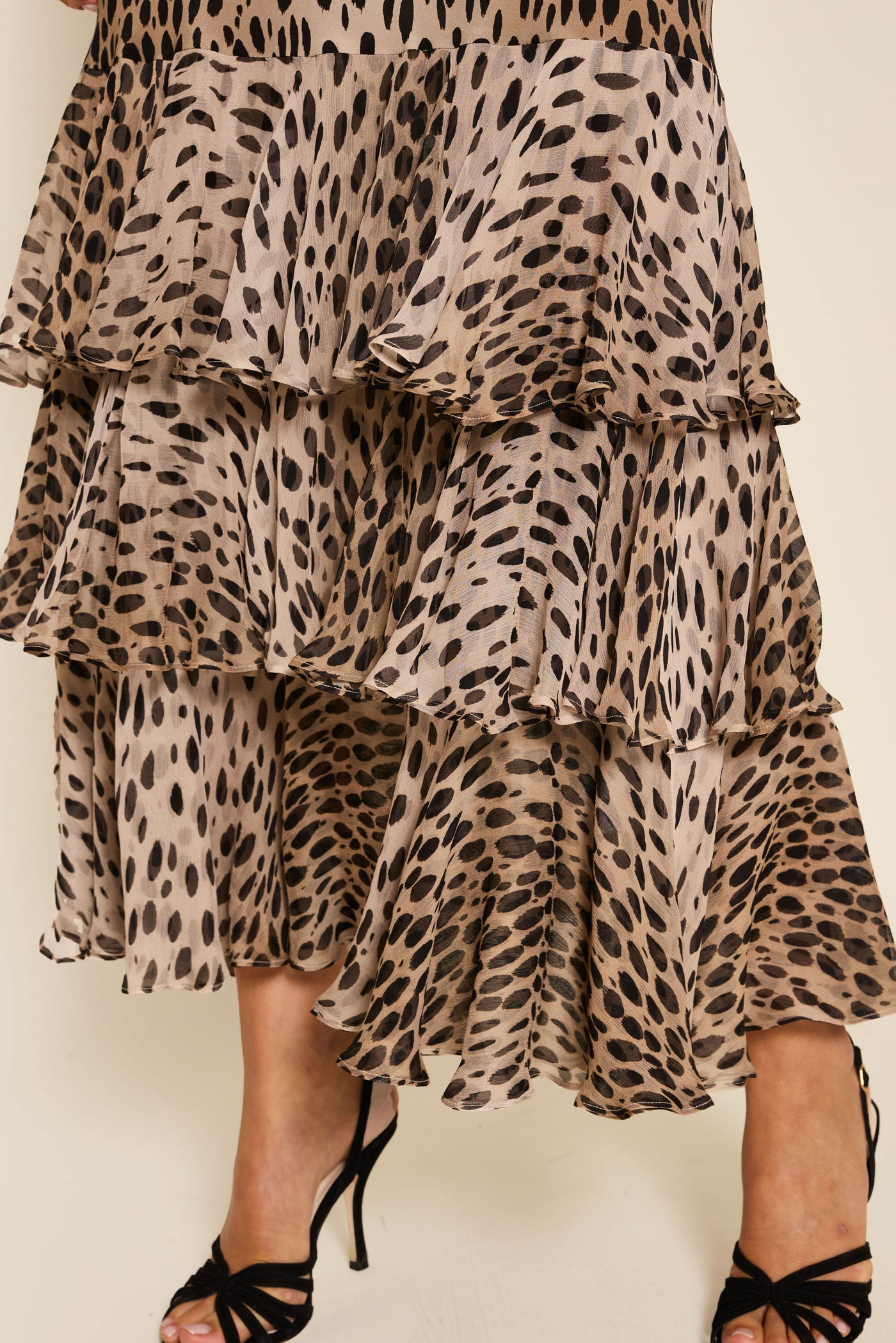 A close-up of the Amelle - Antelope Spot Black tiered ruffled skirt in beige and black animal print silk crepe de chine, paired with black strappy high-heeled sandals.