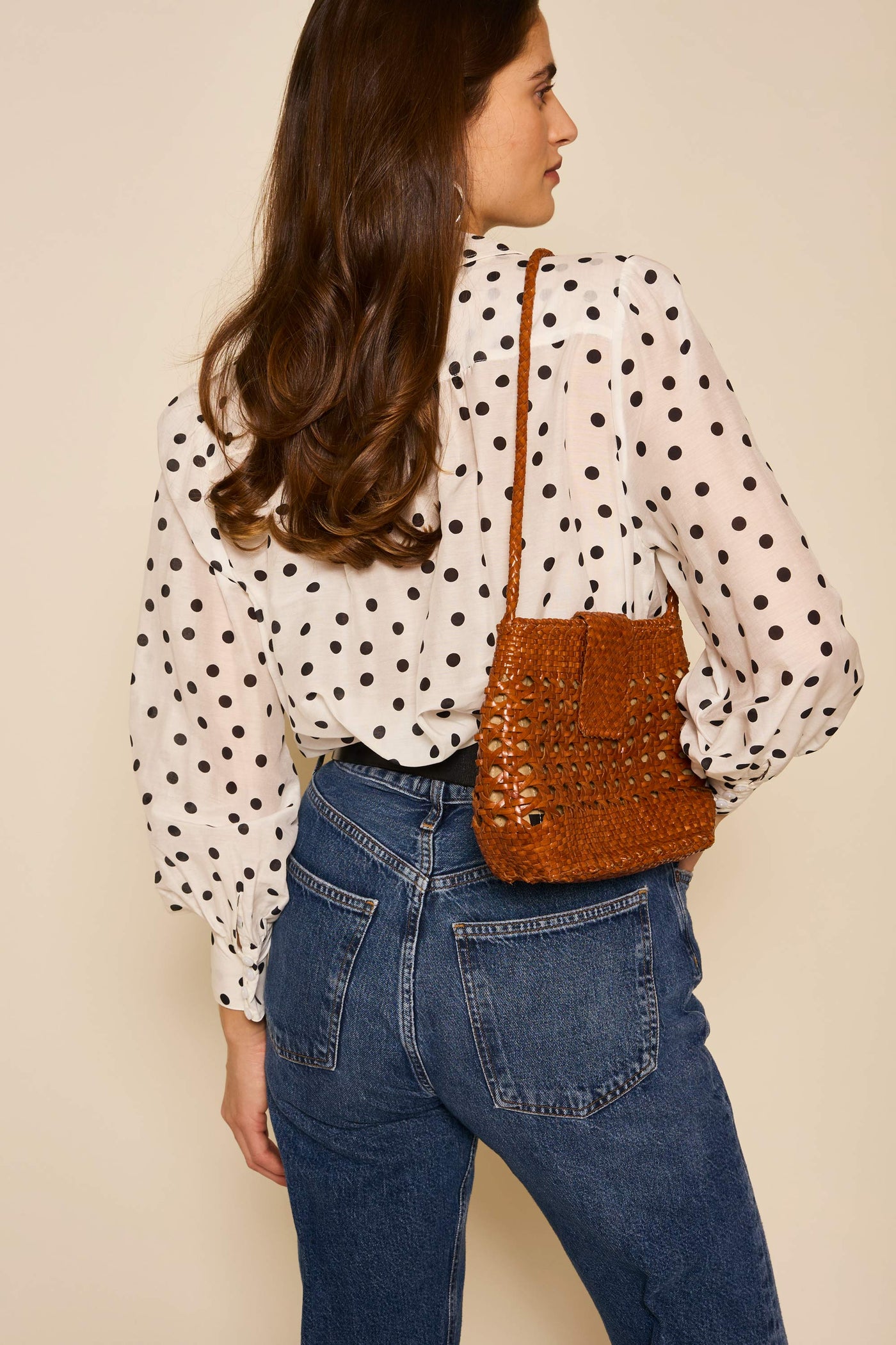 A woman with long brown hair, wearing the Blake - Bohemia Spot Top and blue jeans, stands with her back to the camera. She carries a small brown woven shoulder bag over her shoulder.