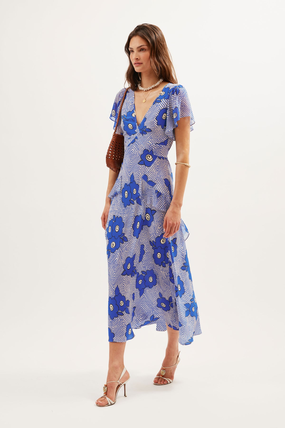 A woman wears the Evie - Milos Floral Blue dress, featuring blue and white florals, with beige heeled sandals, gold jewelry, and a brown handbag, standing against a plain white background.