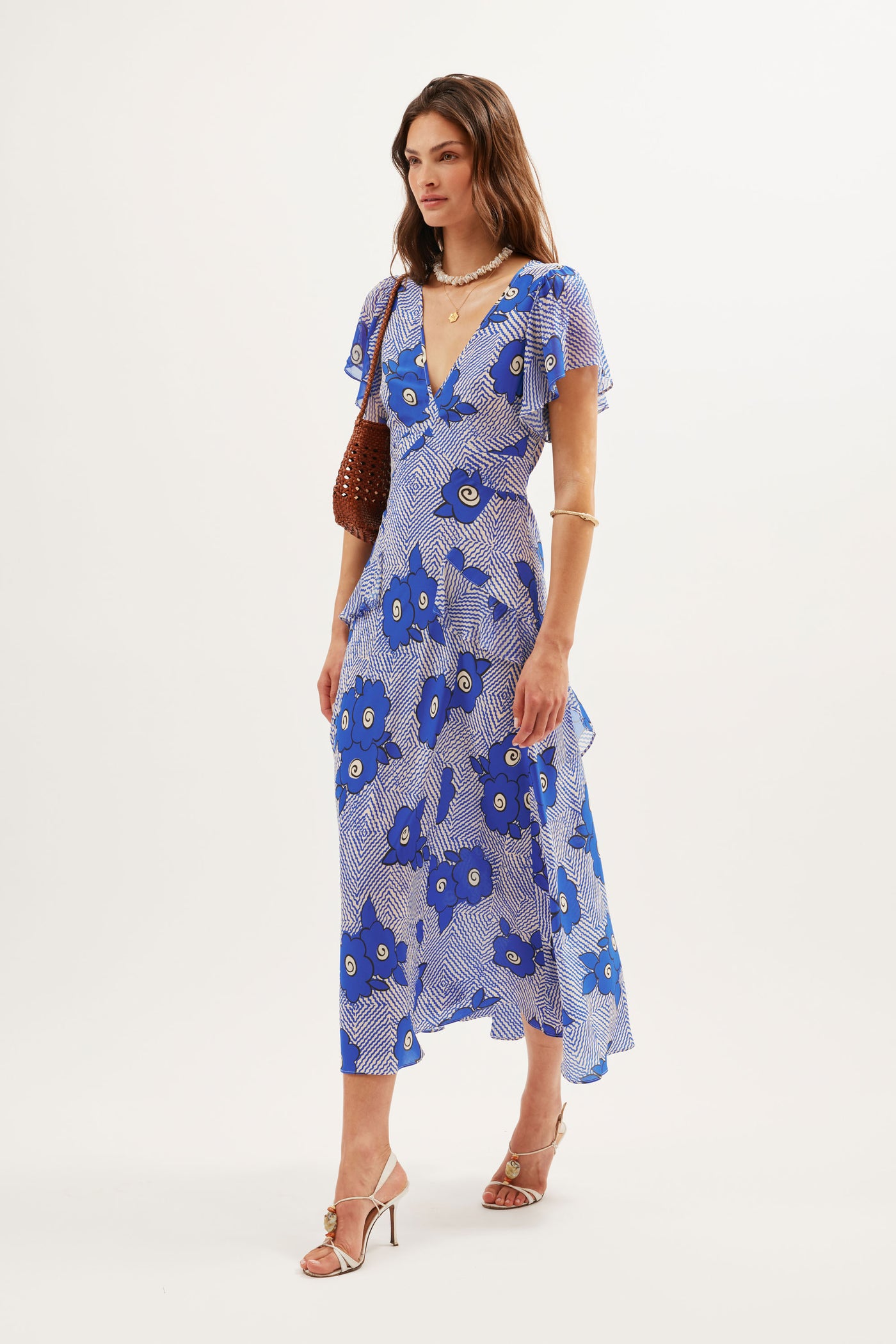 A woman wears the Evie - Milos Floral Blue dress, featuring blue and white florals, with beige heeled sandals, gold jewelry, and a brown handbag, standing against a plain white background.