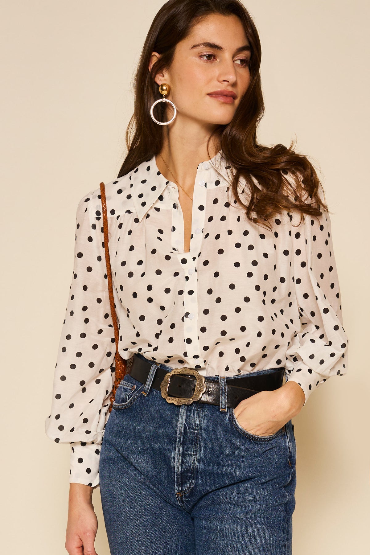 A woman wearing the Blake - Bohemia Spot Top, blue jeans, a black belt with a large buckle, hoop earrings, and a brown shoulder bag stands against a beige background.