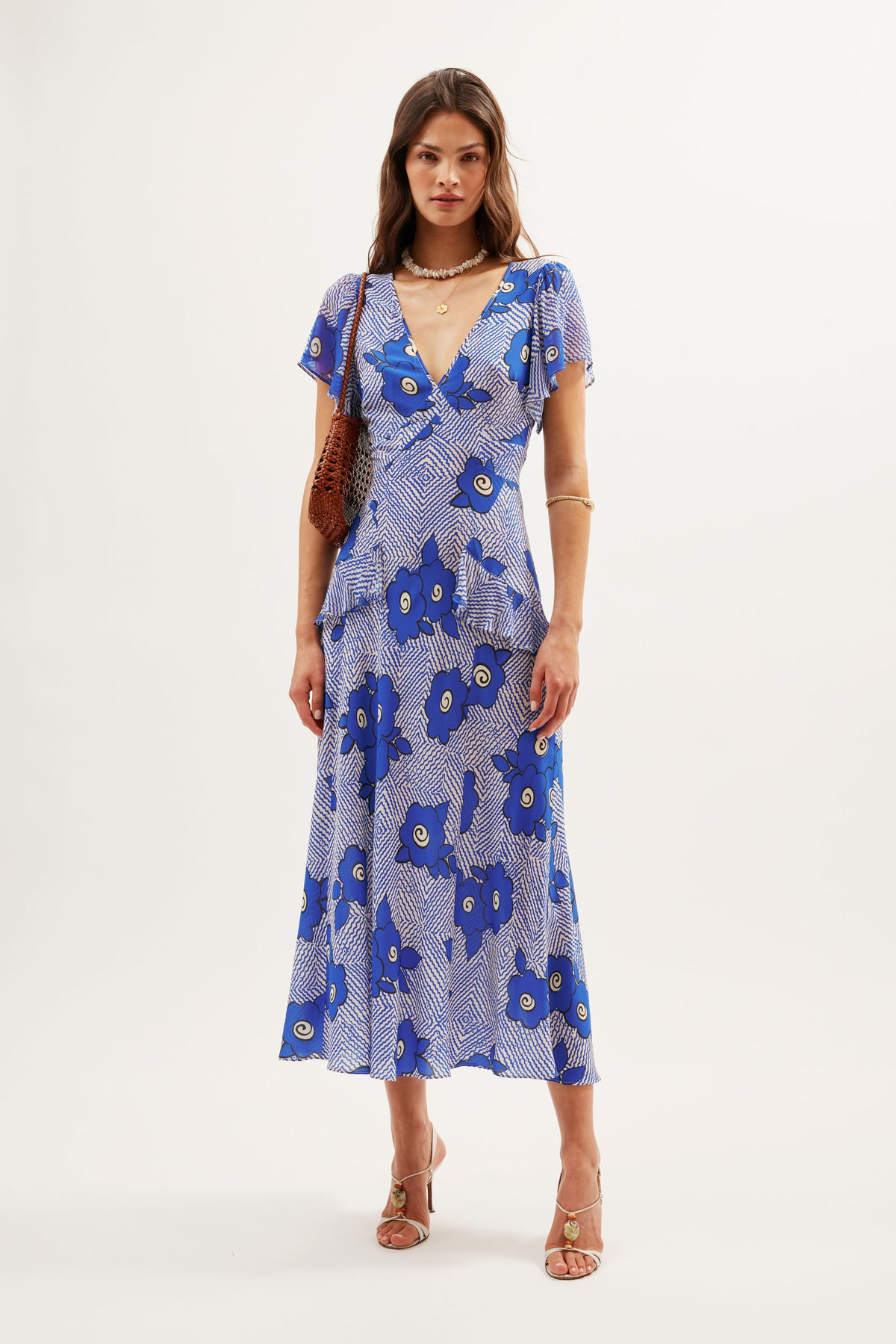 A woman stands in a studio wearing the Evie - Milos Floral Blue dress, a silk crepe de chine midi with bold blue and white florals, short sleeves, and a V-neckline. She pairs it with a brown shoulder bag, gold jewelry, and beige heeled sandals.