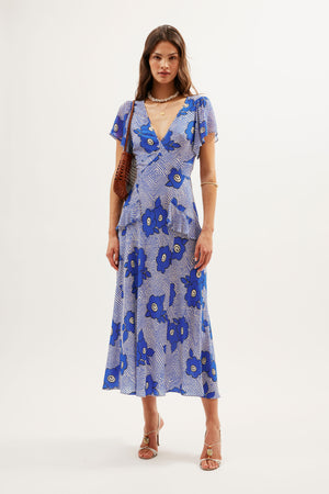 A woman stands in a studio wearing the Evie - Milos Floral Blue dress, a silk crepe de chine midi with bold blue and white florals, short sleeves, and a V-neckline. She pairs it with a brown shoulder bag, gold jewelry, and beige heeled sandals.