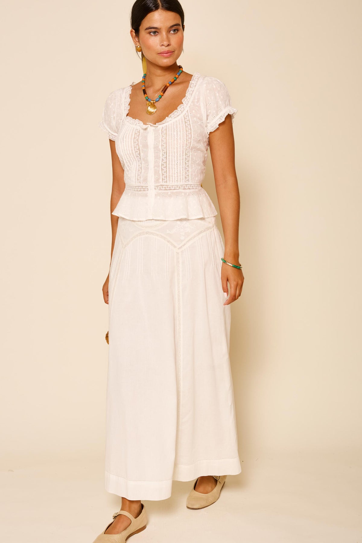 A woman stands against a beige background wearing the Vienne - Pansy Embroidery Ivory, styled with colorful necklaces, a bracelet, and beige flat shoes.