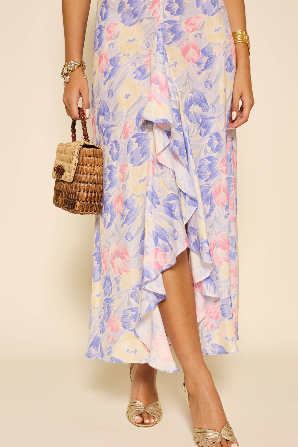 A woman, visible from the waist down against a neutral backdrop, wears the Ariel - Tulip Fields Blue pastel floral ruffled skirt, paired with gold peep-toe heels, gold jewelry, and a woven basket handbag.