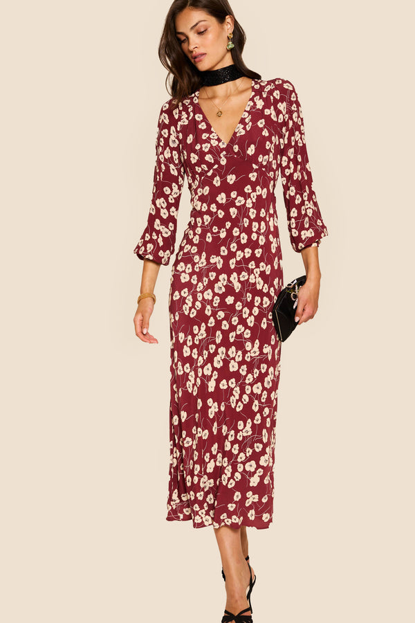 A woman stands against a beige background in the Mavis - Autumn Cherry Maroon dress, a maroon floral-print V-neck midi with long sleeves, styled with a black choker, clutch, and strappy heels—an elegant take on everyday florals.