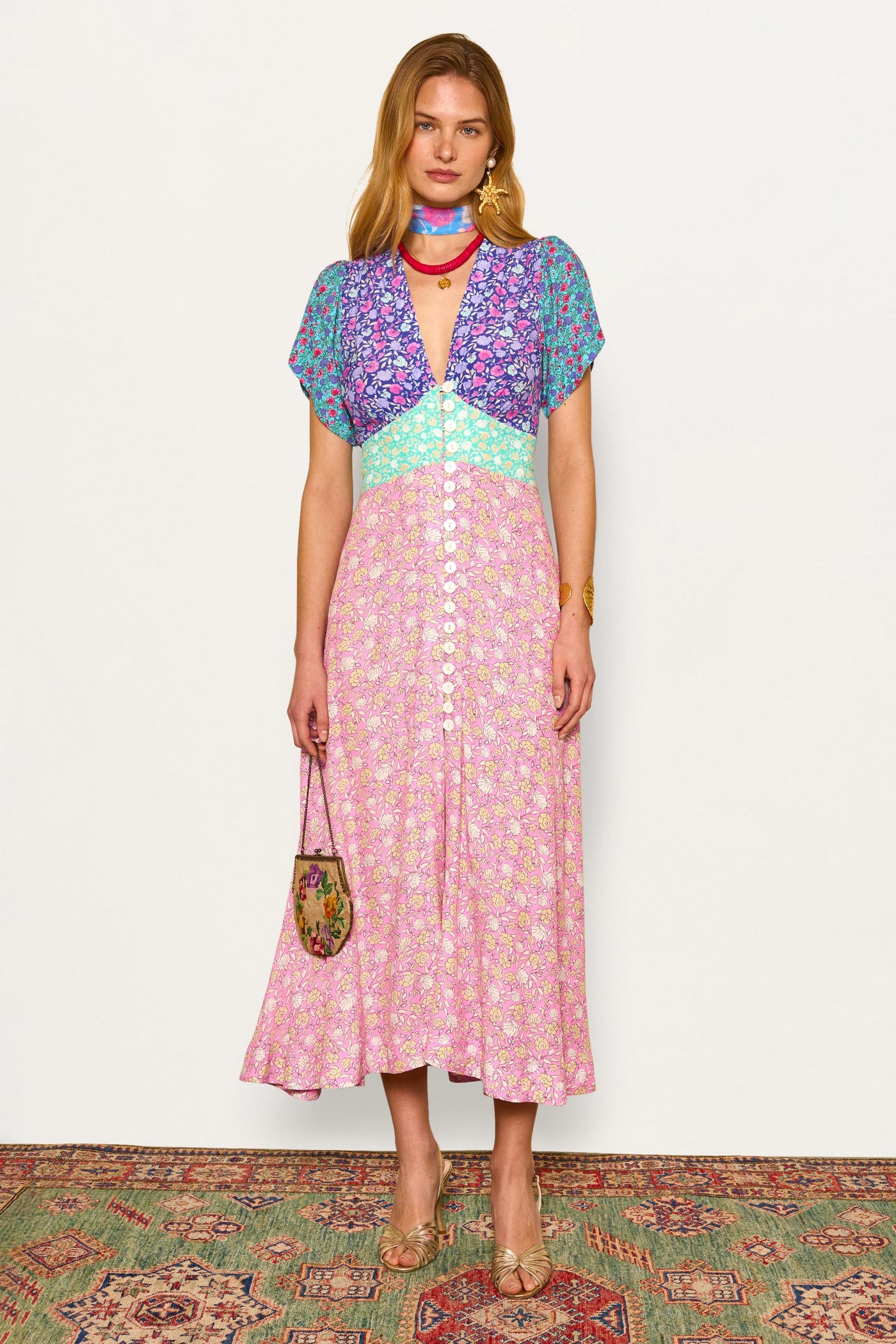 A woman stands on a patterned rug, facing forward against a white wall, wearing the Novara dress in Shell Pansy Mix Multi with short, curved sleeves and a V-neck. She holds a small purse and wears light brown heels and a matching scarf.
