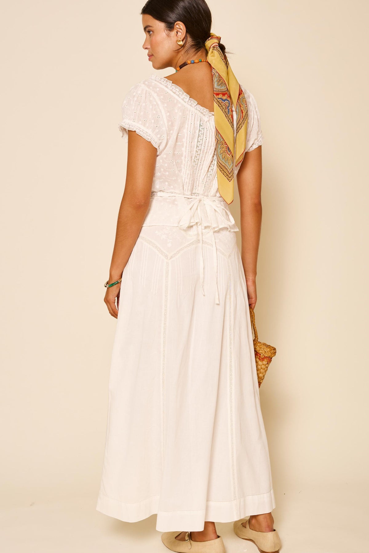 A woman, facing away against a beige background, showcases the Vienne – Pansy Embroidery Ivory dress with lace details and a back bow. She accessorizes with a yellow patterned hair scarf, neutral shoes, and carries a woven bag.