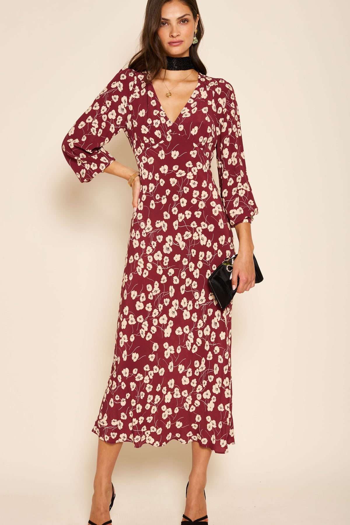 A woman wearing the Mavis - Autumn Cherry Maroon dress—a burgundy midi with floral print, V-neckline, and three-quarter sleeves—holds a black clutch against a light beige background.