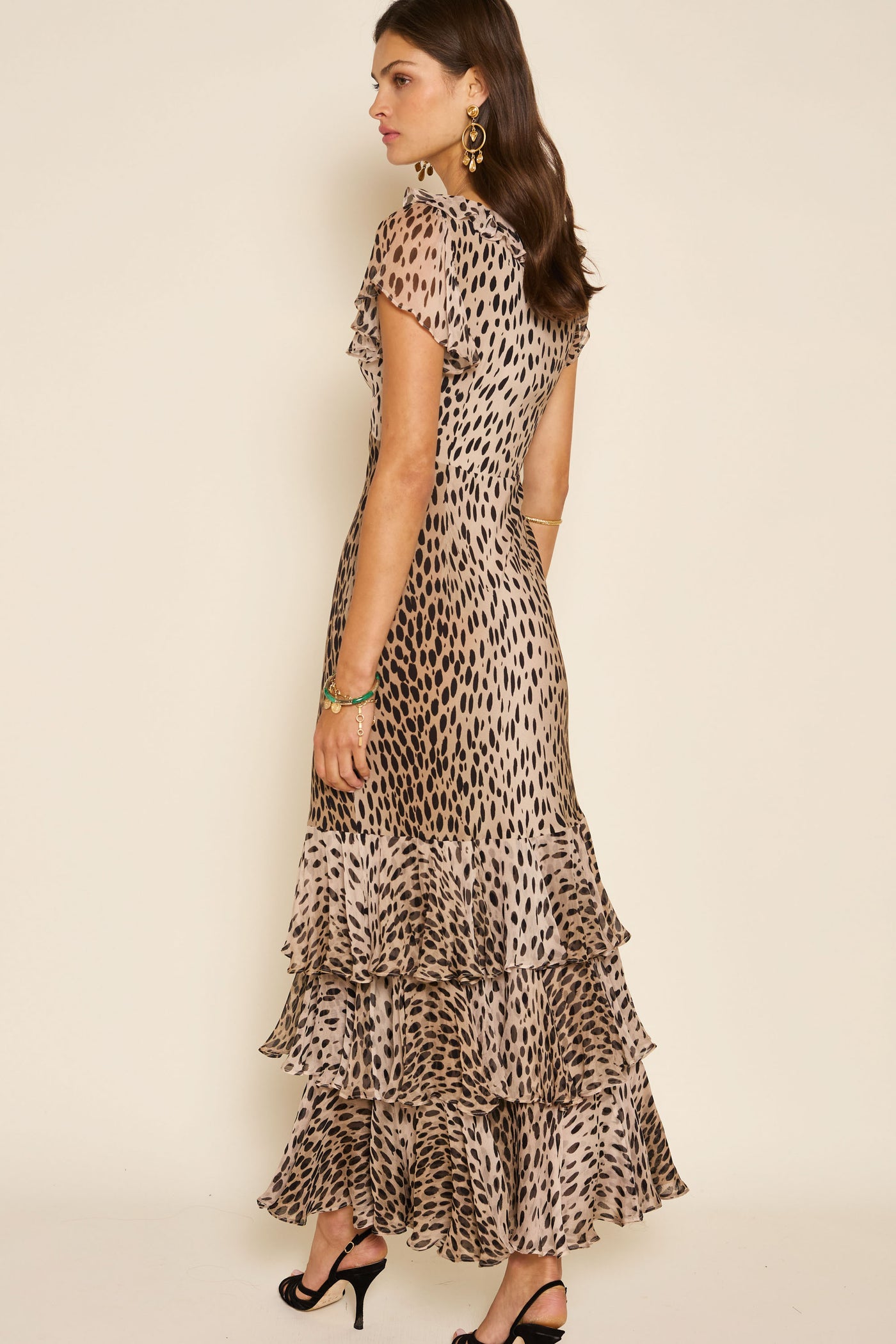A woman stands sideways against a plain background, wearing the Amelle - Antelope Spot Black maxi dress with fluted sleeves, tiered ruffled layers, and black high-heeled sandals.