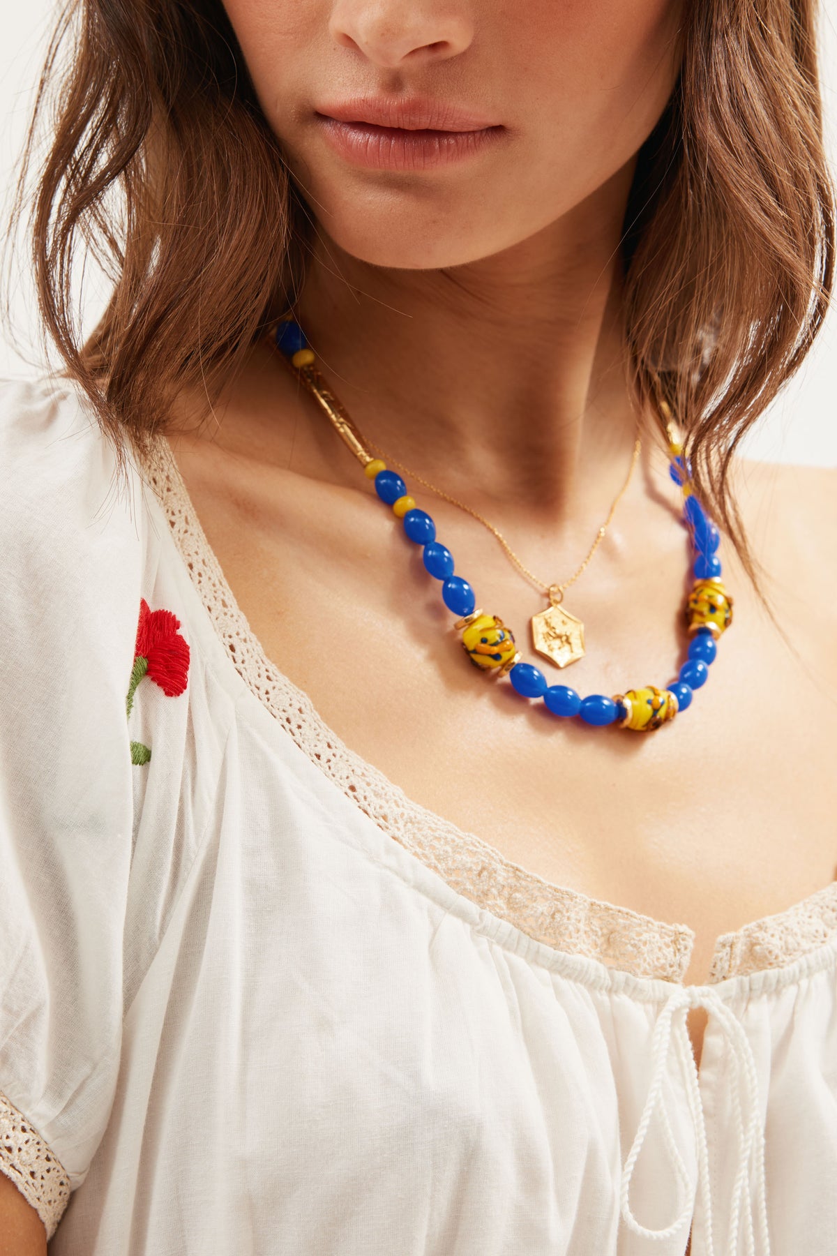 A woman wears a white blouse with lace trim and a red embroidered flower, layered with gold-plated bead necklaces—one being the Denali - Multi necklace, another featuring blue beads, along with a vintage Star of David pendant.