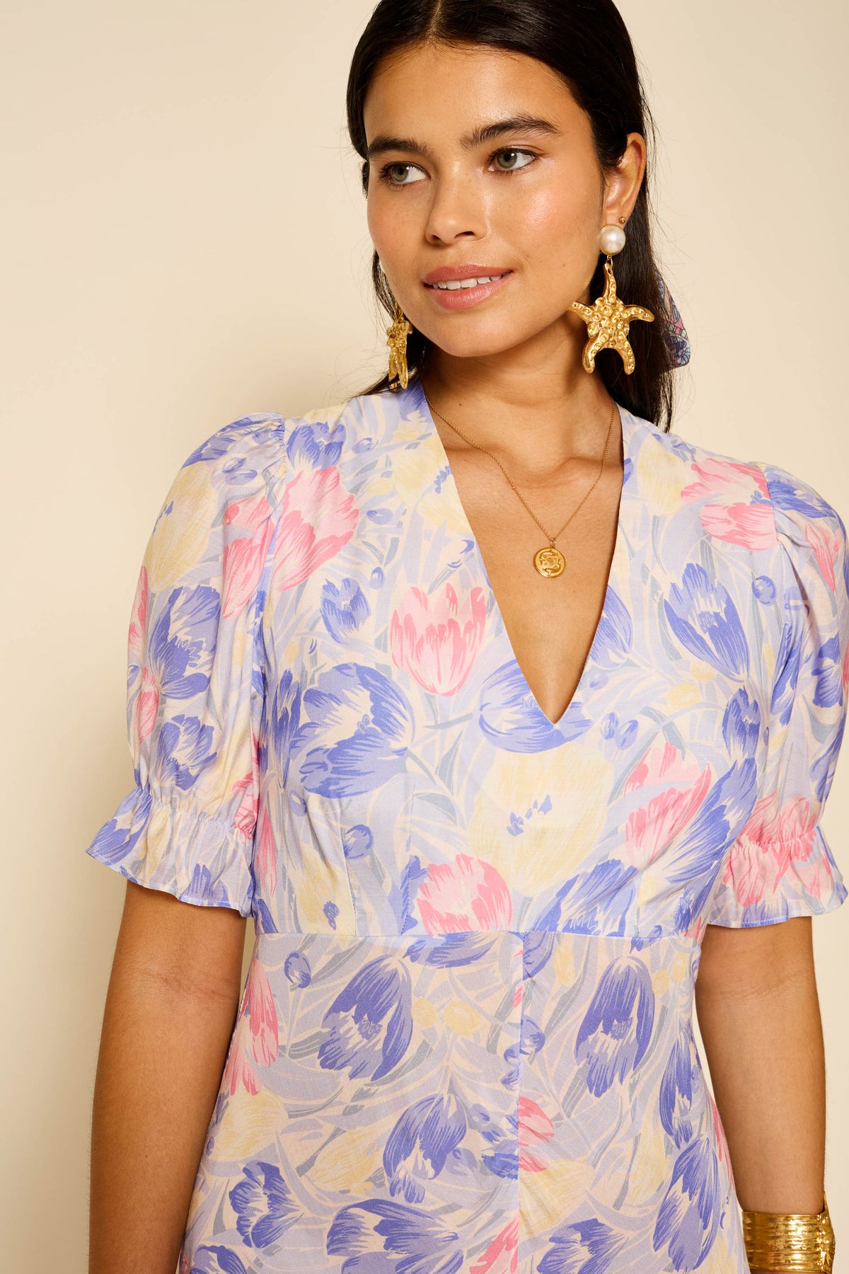 A woman with long dark hair wears Ariel - Tulip Fields Blue, a vintage-inspired dress in soft blue with a V-neck and puffed short sleeves, perfect for special occasions, styled with gold jewelry as she smiles slightly against a neutral background.