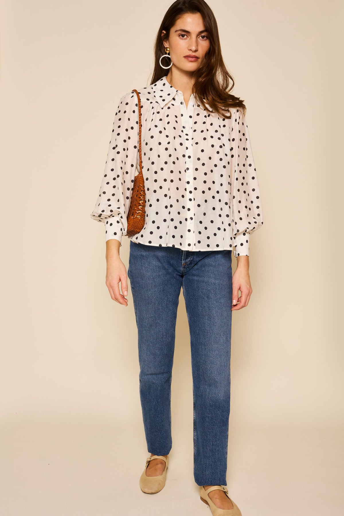 A woman stands against a beige background wearing the Blake - Bohemia Spot Top with black polka dots and an exaggerated collar, blue jeans, beige flats, large hoop earrings, and carrying a brown shoulder bag.